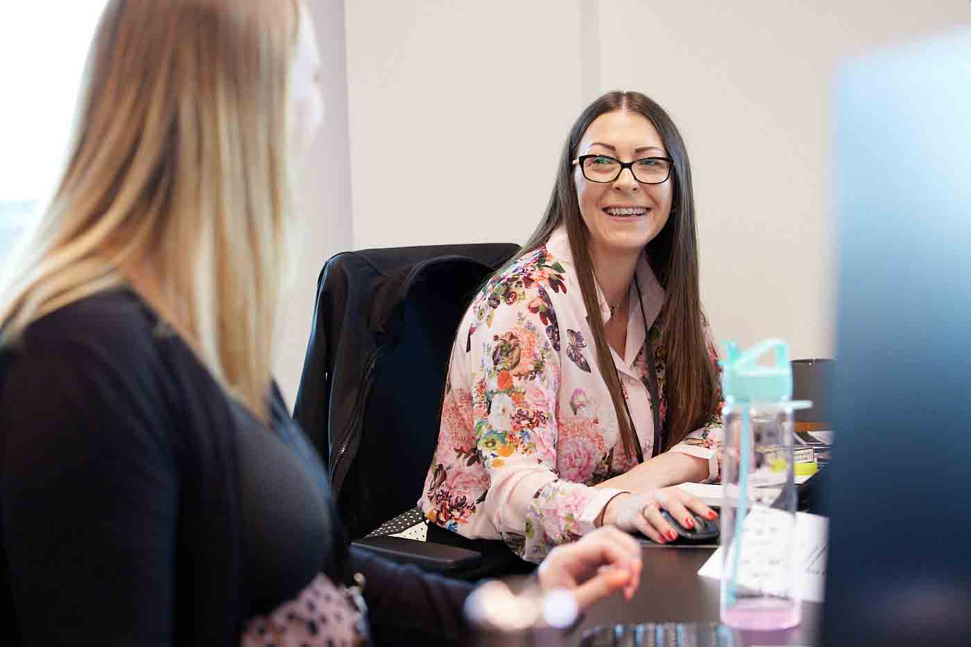 Two women sat at desk looking at each other.
